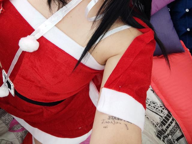 Kkatalina in a red Santa dress with white trim, showing her tits in a white bra, black hair, arm tattoo, and red sleeve, on a bed with colorful pillows.