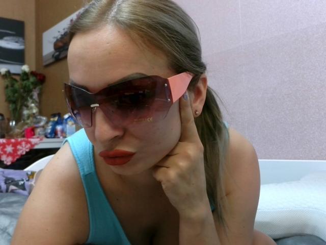 AngelikaStar69 with pink-tinted sunglasses, red lipstick, blue tank top, and hand on cheek, showing her cleavage, has light brown hair in a ponytail, and is wearing a small earring.