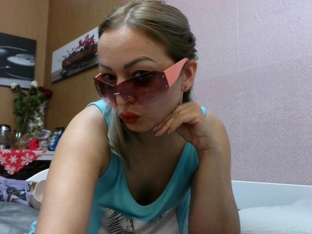 AngelikaStar69 with blonde hair, pink-tinted sunglasses, red lipstick, and a blue tank top, leaning on a bed with her hand under her chin, showing her tits.