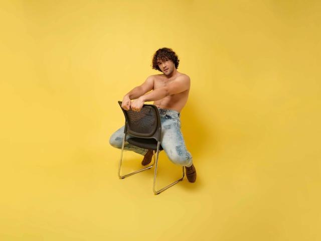 Shirtless FreyrStyle69 with curly brown hair, muscular build, and light skin, sitting on a black mesh chair, wearing loose blue jeans and brown boots, against a yellow background.
