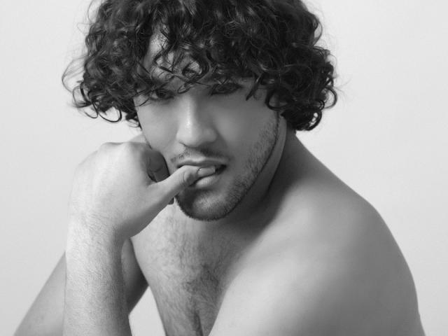 FreyrStyle69 with curly dark hair, biting finger, shirtless, showing a hairy chest, and a sexy, handsome face, in a black-and-white photo.