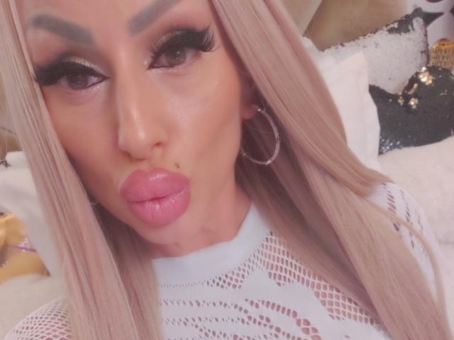 HotBebe69x mit pinkem Lippenstift, vollen Lippen und einer kleinen Sommersprosse auf der Nase. Sie hat langes blondes Haar, dichte Wimpern und trägt ein weißes Mesh-Oberteil..