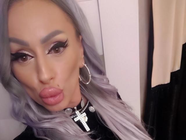 HotBebe69x avec des lèvres pulpeuses, des cheveux gris ondulés, des sourcils épais, de longs cils, un fard à paupières rose, un collier avec une croix en argent et de grandes boucles d'oreilles. Elle fait une moue..