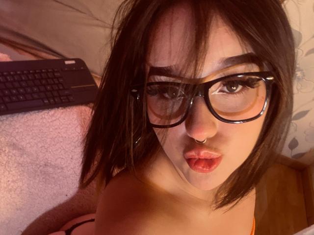 CristySanz with dark brown hair, wearing glasses and a nose ring, pouting with red lips, looking up at the camera, laptop in the background, lying on a pink blanket.