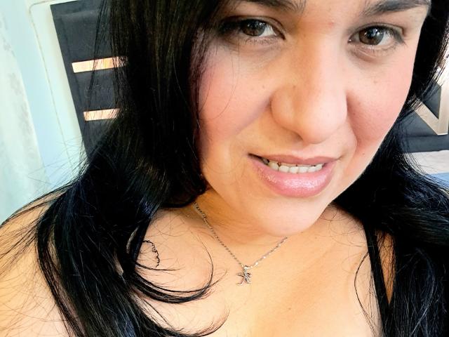 VivianaThompson has dark black hair, brown eyes, and a slight smile. She's wearing a silver necklace with a small spider design, showing her bare shoulders and shiny lips.