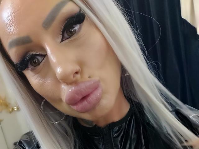 HotBebe69x con piel brillante, labios rosados y gruesos, cabello blanco y negro, cejas pobladas, pestañas largas y un piercing en la nariz, mirando directamente a la cámara con un ligero bostezo..