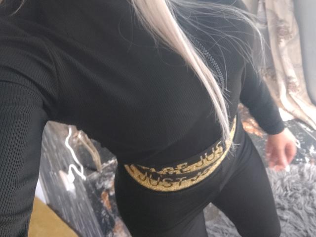 HotBebe69x portant un haut noir à manches longues et côtelé, un pantalon noir et une ceinture dorée avec le mot "MUSHROOM" en lettres capitales. Cheveux longs et blancs, veste noire zippée, debout légèrement..