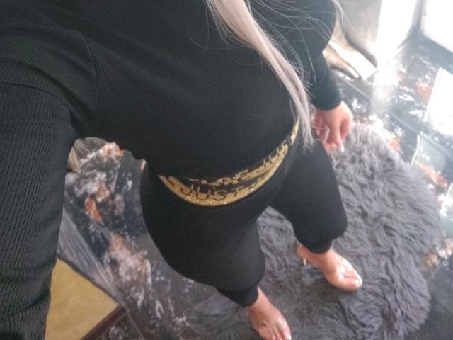 HotBebe69x in black ribbed top, black pants, gold belt, bare feet, standing on dark textured rug, long silver hair, selfie angle, upper body and legs visible.