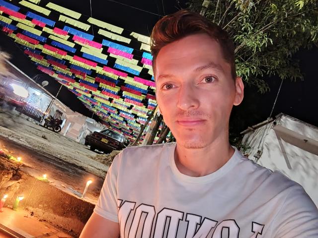 AlanPussy with short brown hair, slight mustache, white shirt, standing in front of colorful string lights at night, looking directly at camera with a neutral expression.