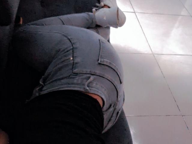 JackelineRoset's achterkant in strakke, blauwe jeans, licht gescheurd op de bil, met een zwart shirt, liggend op een donkere bank, met witte sneakers aan, op een glanzende betegelde vloer..