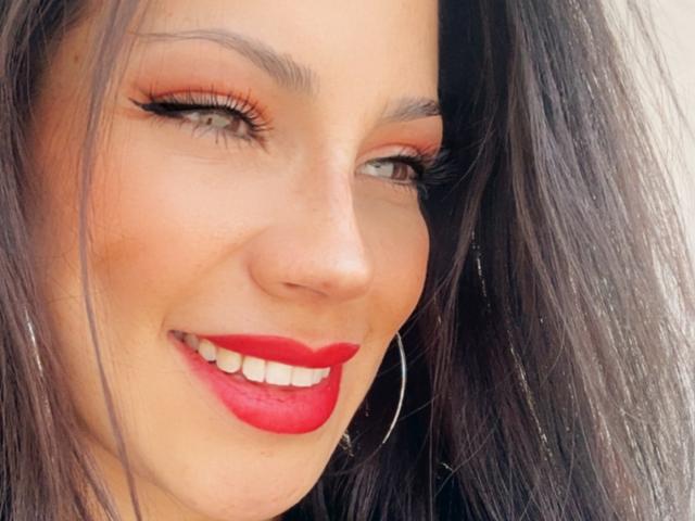 Liliysweet's face, showing her red lipstick, black eyeliner, hoop earrings, and smiling with her green eyes, brown hair, and fair skin. Her red lips are her biggest feature.
