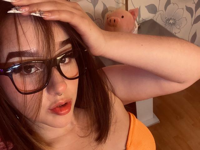 CristySanz with glasses, nose ring, and orange top, hand in brown hair, looking at camera, mouth slightly open, wooden floor, pink piggy in background.