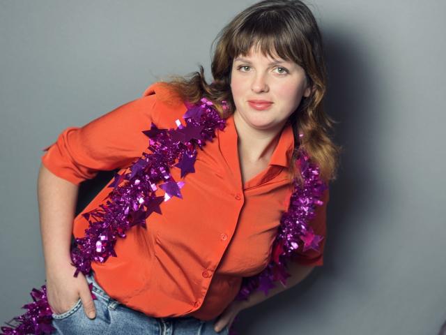 StaceyLex with purple tinsel hanging on her orange shirt, blue jeans, and brown wavy hair, leaning forward with a slight smile, hand in pocket.