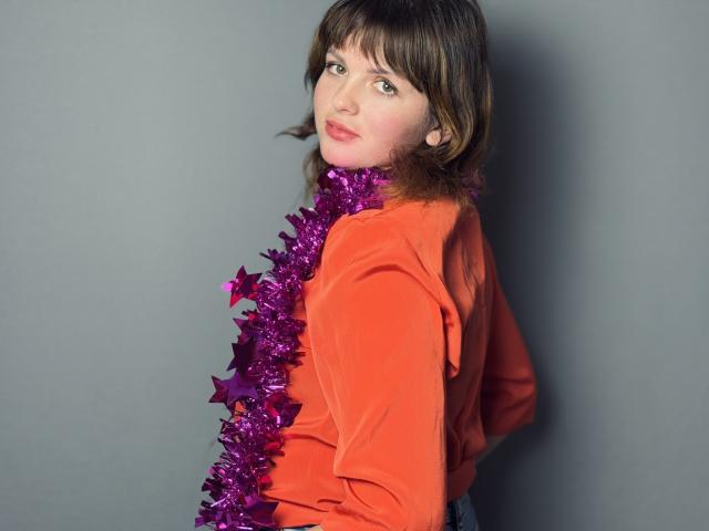 StaceyLex with medium brown hair, wearing an orange shirt, has a purple tinsel garland around her neck, looking back with a slightly surprised expression.