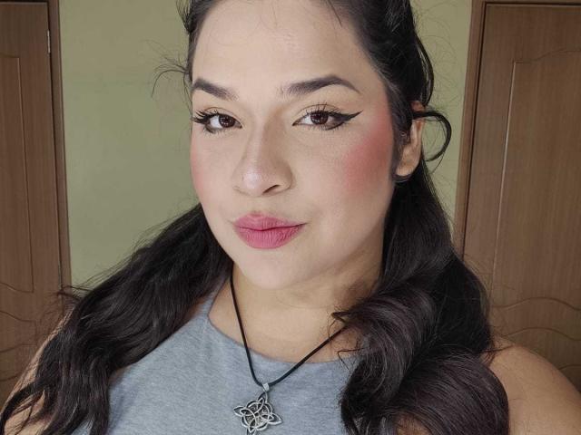 Antonela, avec ses cheveux foncés, ses joues rosées, son eye-liner noir, son rouge à lèvres rose, portant un haut gris et un collier avec un motif de nœud celtique, souriant doucement..