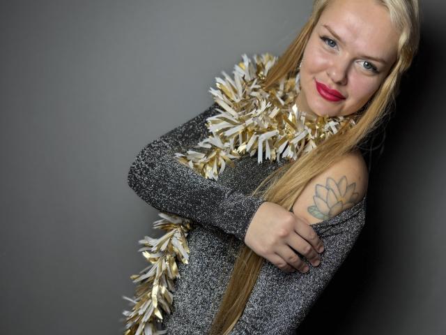 Melissa. Gatas con cabello rubio largo, usando una blusa gris brillante, guirnaldas doradas y blancas, y lápiz labial rojo, sonriendo, con un tatuaje en el hombro derecho, brazos cruzados, fondo gris..