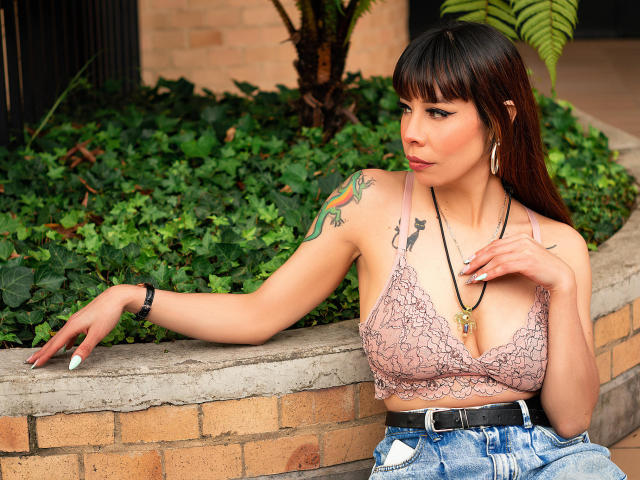 AriiannaHot with brown hair, bangs, and tattoos, wearing a pink lace bra and distressed jeans, leaning on a brick wall, looking to the side, with a necklace.