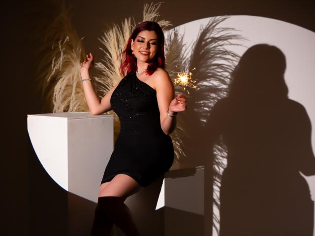 ZoeeMar with red hair, wearing a black, one-shoulder, sparkly dress, holding a sparkler, standing next to a white cube, shadowed against a wall with pampas grass.