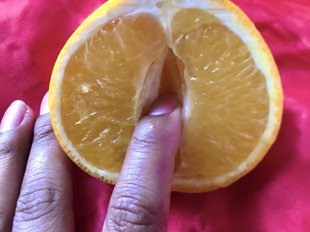 SophieSonyas's finger is in a juicy, halved orange with a red cloth underneath. Her finger is on the orange's middle, pointing at the center.