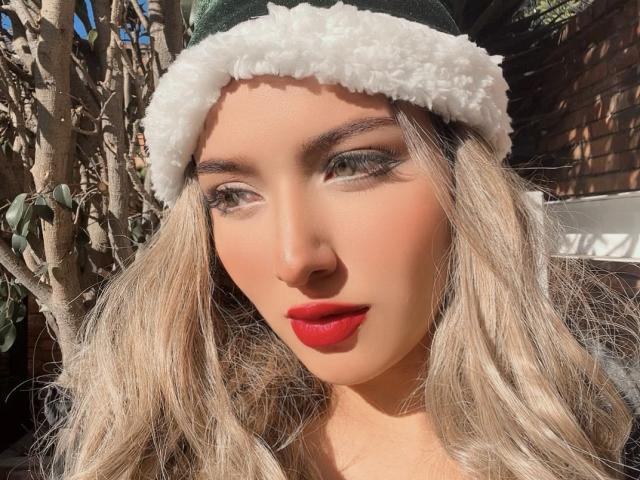 LillyTaylor with wavy blonde hair, green furry hat, red lipstick, and eyeliner, looking to the side in sunlight, showing off her cute ass-face and flirty expression.