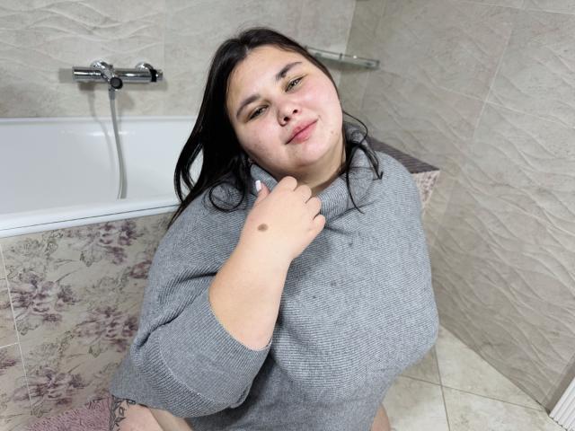 KaidoKun with light skin, black hair, and a cute face, wearing a gray sweater, sitting by a bathtub, holding her shoulder with a small mole on her hand.