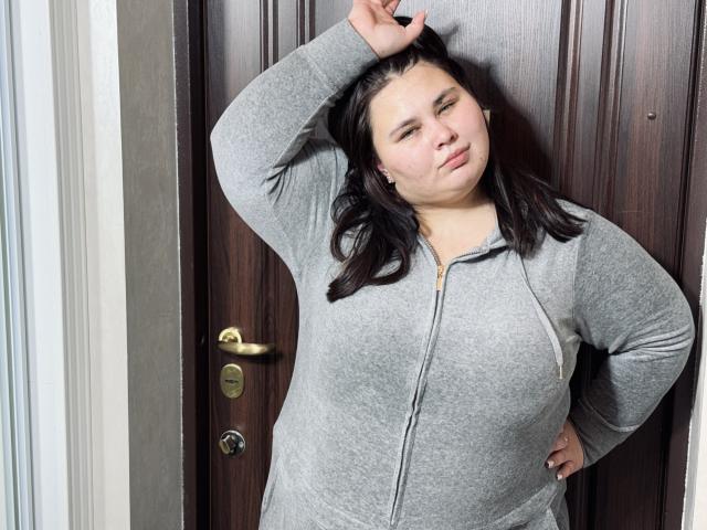 KaidoKun, a chubby woman with dark hair, wearing a gray zip-up hoodie, stands against a wooden door, hand in hair, looking slightly annoyed, with a small nose.