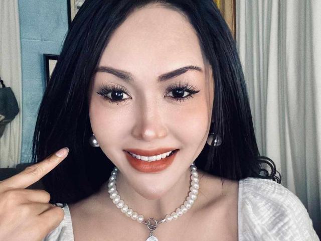 QueenDomCum has black hair, brown eyes, and is smiling with orange lipstick. She's wearing a pearl necklace and earrings, pointing at her lips with a finger.