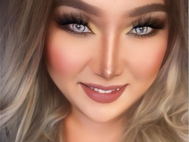 HeavenlyGoddess tem olhos azuis, cílios longos e um rosto sorridente com blush rosa, batom nude e cabelo loiro ondulado. Sua maquiagem inclui delineador escuro e sombra amarela suave..