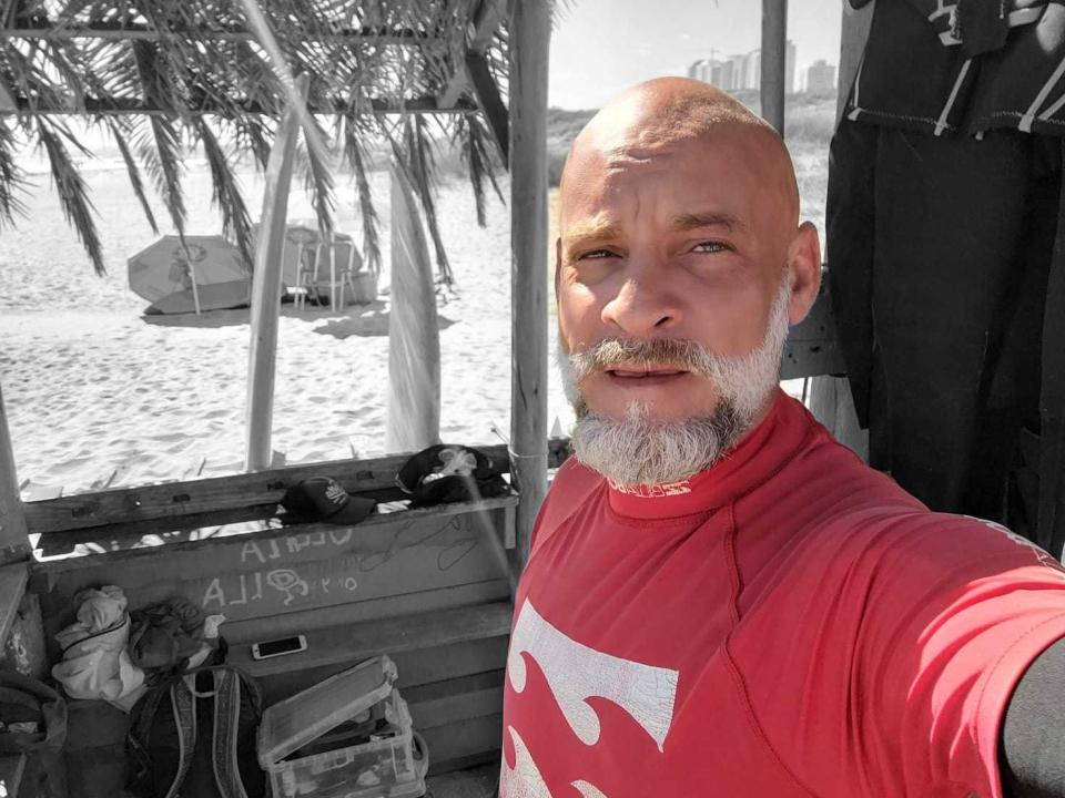 Sergio Weiske, schon bald mit einem weißen Bart, trägt ein rotes Hemd mit einem weißen Logo und steht in der Nähe einer Strandhütte. Er blickt mit einem leichten Lächeln in die Kamera..