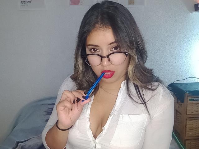 LuluHorn with medium tits, wearing glasses and a white button-up, holding a blue pen to her red-lipped mouth, in a room with a bed and wooden box.