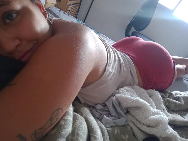 LuluHorn sdraiata su un letto, indossa una canotta bianca e pantaloncini rosa aderenti, mostrando il suo sedere rotondo e il braccio tatuato, e guarda la fotocamera con un leggero sorriso..