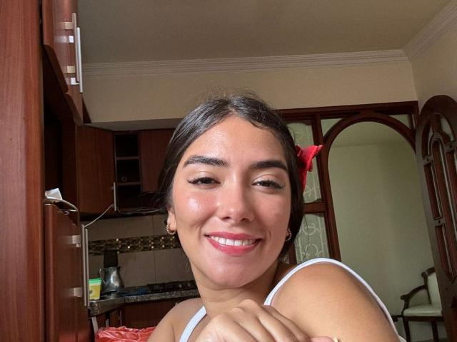 SharonKlart with shiny skin, brown hair, red bow, white strap, smiling, showing teeth, holding arm, brown wooden cabinet, mirror, and kitchen in background.