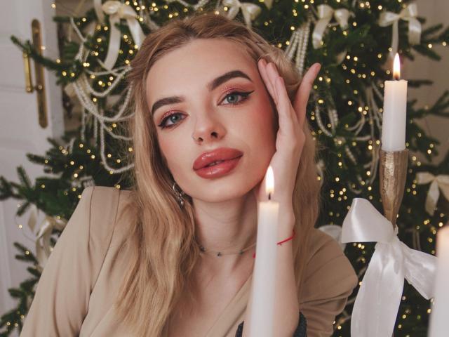 Adele Clifford, avec ses lèvres rouges et pulpeuses, ses cheveux blonds et son eye-liner, touchant son visage, portant un haut beige, devant une bougie allumée et un sapin de Noël..