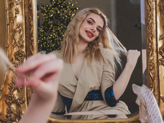 Adele Clifford, con i capelli biondi, il rossetto rosso e le maniche blu ricoperte di paillettes, si tocca i capelli in uno specchio con cornice dorata, indossa un abito beige e sorride. Una mano tiene un pennello..