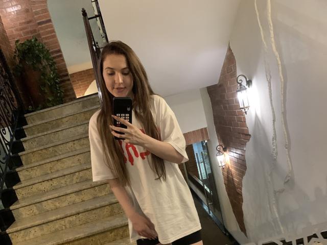 CaramellSex with long brown hair, white shirt, and red logo, taking a mirror selfie with phone in stairway, holding her pussy with right hand.