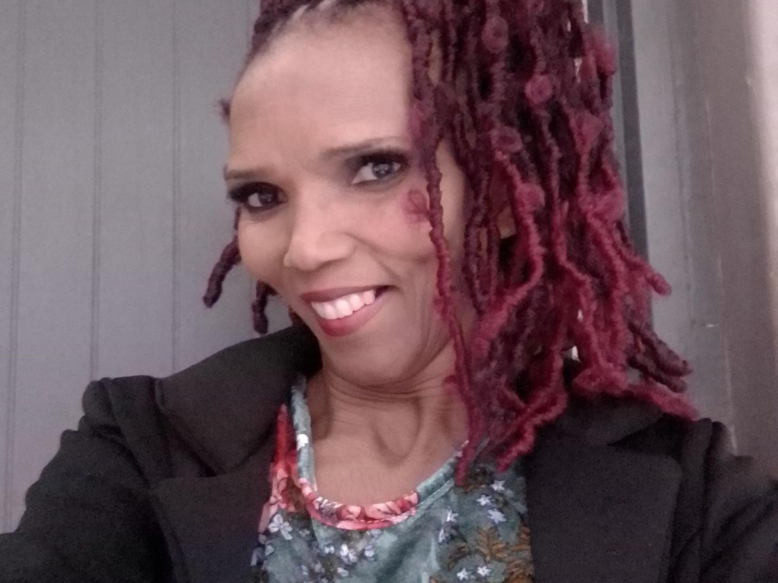 ASweetCherryFace with maroon dreadlocks, smiling with white teeth, wearing a black jacket and floral shirt, has a small cut on the right cheek, standing in front of a grey wall.