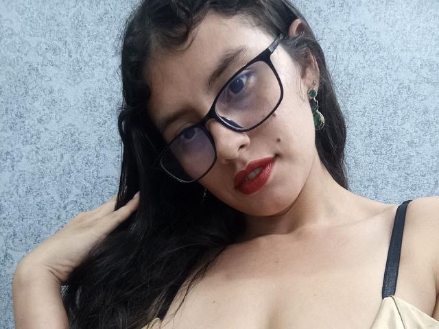 MadissonFontaine with dark wavy hair, glasses, red lipstick, green earrings, and a beige top showing black straps, hand in hair, looking at camera, showing her tits.