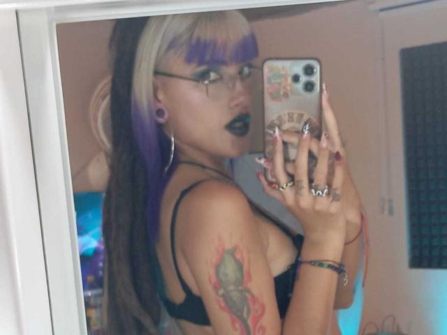 SharlotteX, com cabelo roxo e loiro, batom preto, óculos, sutiã preto, tatuagem no braço, segurando o celular em uma selfie no espelho, mostrando os seios e alargadores de orelha..