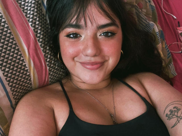 VictoriaSaens with dark hair and bangs, smiling with black eyeliner, wearing a black tank top, gold hoop earrings, and a necklace, lying on a patterned pillow, showing a tattoo.