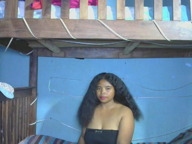 RineeAimee with dark curly hair, brown skin, and small boobs, wearing a black strapless top, standing in front of a wooden bunk bed with colorful bedding above.