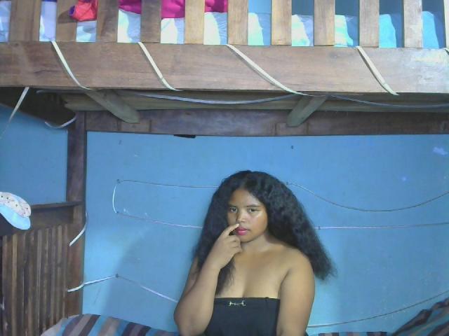 RineeAimee has dark curly hair, brown skin, and is wearing a strapless black top, with her finger touching her red-lipsticked mouth, standing in front of a wooden loft.