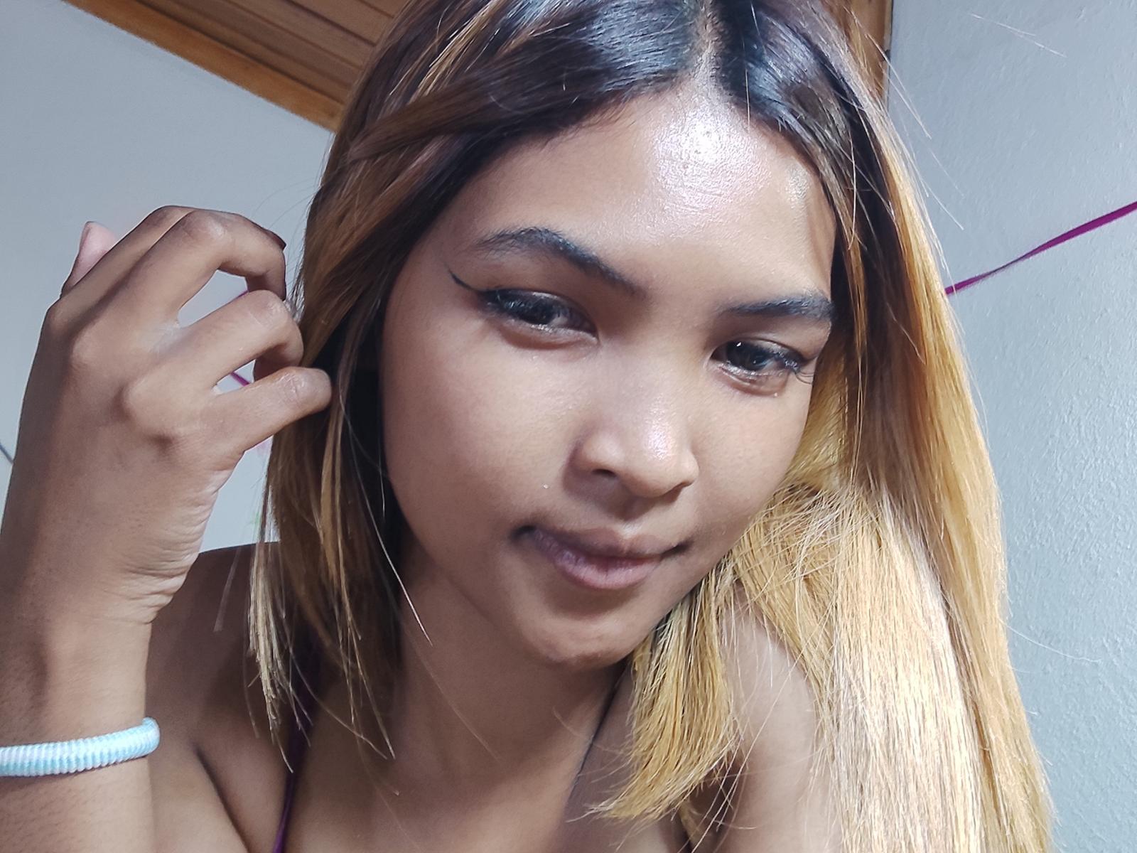 NinahMe tem a pele marrom escura, cabelo loiro liso com mechas, e está sorrindo com a mão perto da orelha. Ela está usando uma pulseira branca e tem delineador preto estilo "asa de borboleta"..