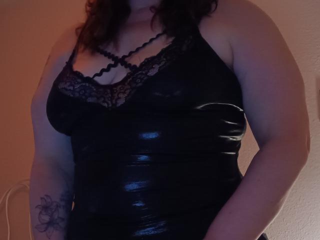 Fecha: Una mujer con curvas, vestida con una blusa de encaje y cuero negro, mostrando un busto grande, tatuaje en el brazo derecho, cabello oscuro ondulado, iluminación tenue, luz suave sobre la piel..