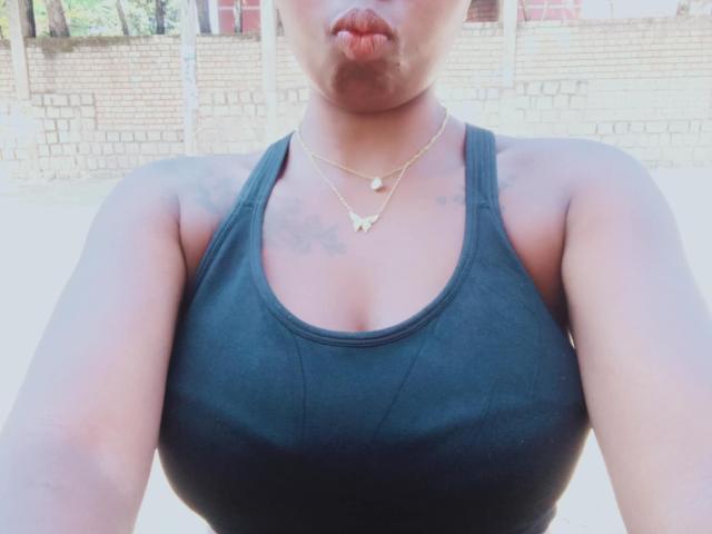 Jobba has dark tits in a black tank top, puffy lips, tattoos on her shoulders, and two gold necklaces. Her skin is dark, and her arms are in the frame.