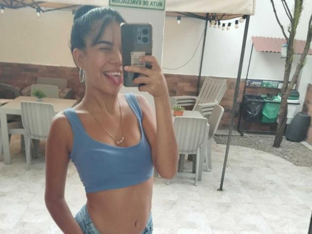 MarySmiith, con cabello oscuro y corto, sacando la lengua, vistiendo un top deportivo azul y jeans, mostrando sus pequeños pechos y glúteos planos, tomando una selfie frente al espejo..