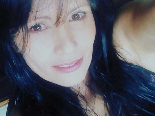 DulceHotShary has dark blue, wavy hair, brown eyes, and a pinkish lip. She's smiling with her tits partially visible in the right corner.
