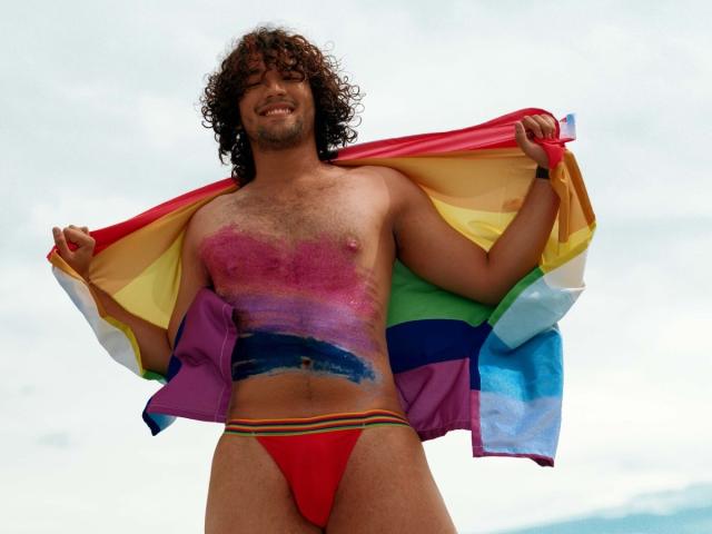 FreyrStyle69 with curly brown hair, hairy chest, rainbow flag painted on torso, red speedo, holding flag, smiling, standing outdoors, visible cock in speedo.