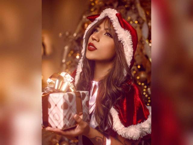 CourtneyLow with long brown hair, wearing a red velvet Santa cape and hat, holds a gift with a golden ribbon, looking up with red lipstick. Warm, blurred background.