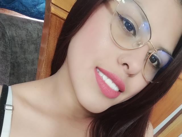 ElaiaaBakerr with pink lipstick, white teeth, glasses, nose piercing, and brown hair, showing her face and neck in a selfie, wearing a white top with black straps.