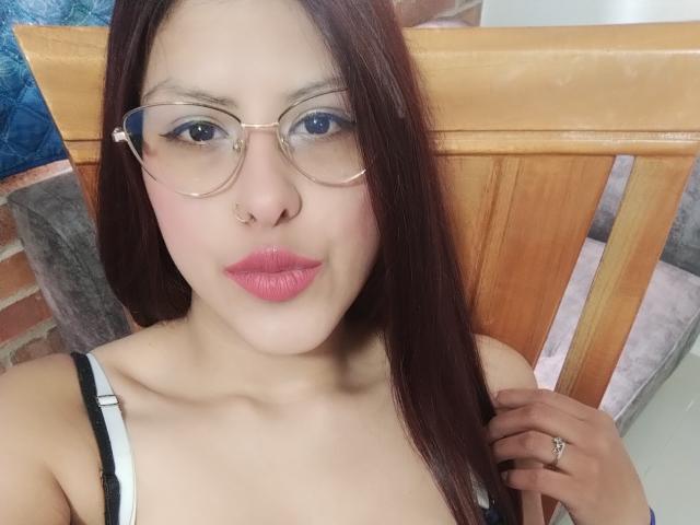 ElaiaaBakerr with pink lipstick, glasses, nose ring, and brown hair, holding her tank top strap, showing her small tits, sitting in a wooden chair.